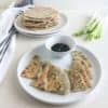 Wedges of scallion pancakes on round white plate with small white bowl of dipping sauce, next to white kitchen towel with gray stripes, stack of pancakes on round white plate next to green onions in the background