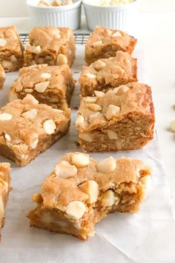 blondie bars on parchment paper one with bite taken out, macadamia nuts next to blondies, ramekins of white chocolate chips and macadamia nuts in the background