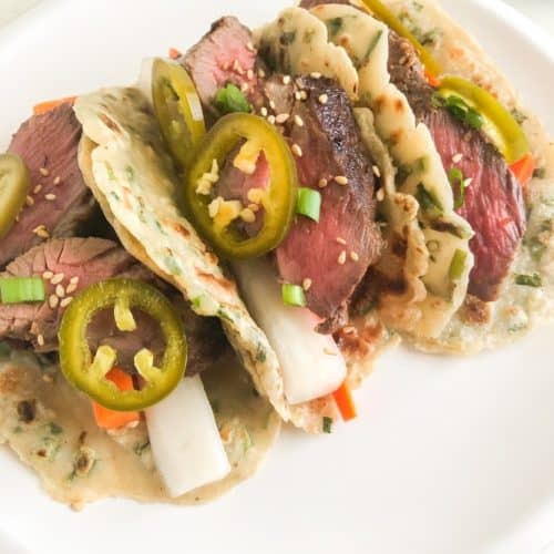 Scallion pancake tacos on round white plate. Tacos filled with sliced short ribs, pickled daikon, carrot, and jalapenos.