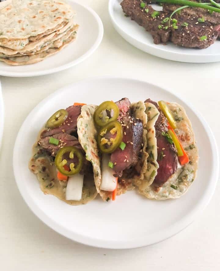 3 scallion pancake tacos on round white plate. Tacos filled with sliced short ribs, pickled daikon, carrot, and jalapenos. Small round white plates in the back ground, one with stack of scallion pancakes and the other with Korean short ribs topped with green onions.