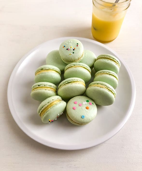 Green lime curd macarons on medium round cream plate next to mason jar with lime curd and silver spoon.