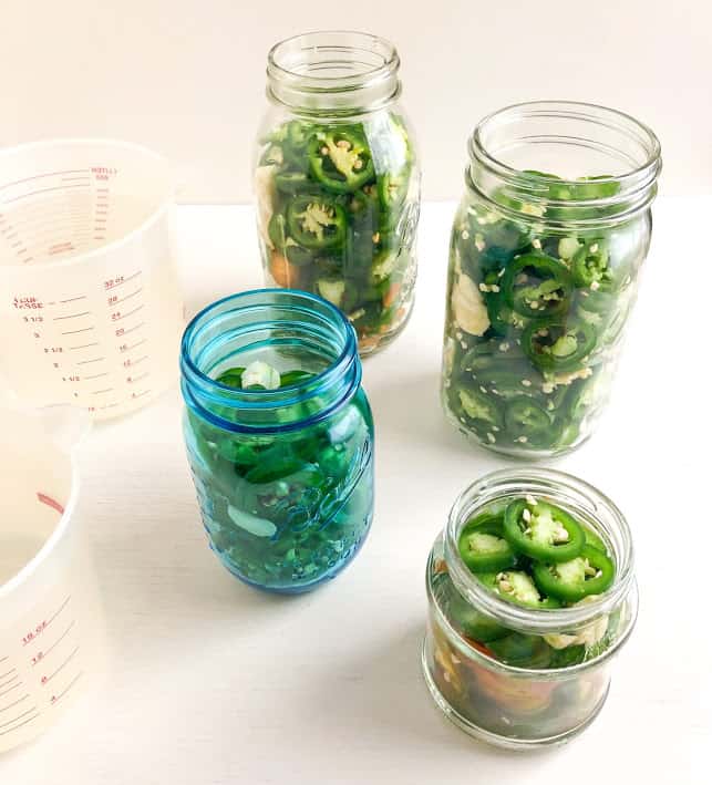 Sliced jalapenos and smashed garlic in jars, pickling liquid in measuring cups.