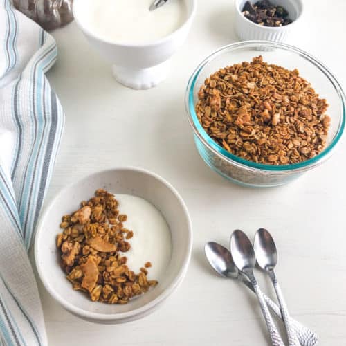 Small round bowl with yogurt and topped with granola. White kitchen towel with gray and blue stripes and silver teaspoons next to bowl. Round glass container with granola, white footed bowl with yogurt, small round ramekin with chocolate chunks, and silver pumpkin in the back ground.