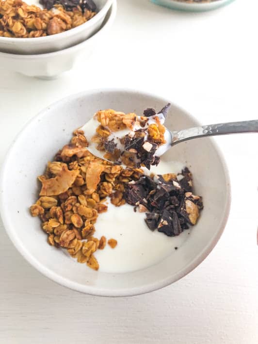 Small round cream bowl with small speckles filled with yogurt, granola, and chocolate chunks. Small silver teaspoon with scoop of yogurt, granola, and chocolate.  Additional bowl of granola in the backround.