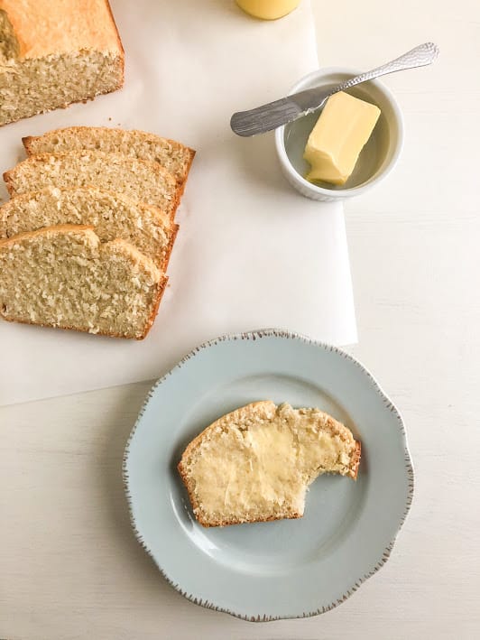 Slice of coconut bread on round light blue plate with butter on top with bite taken out of the slice. Sliced bread and the remaining loaf on parchment paper next to a small white round ramekin with butter and a silver butter knife. Glass jar with lime curd in the background.