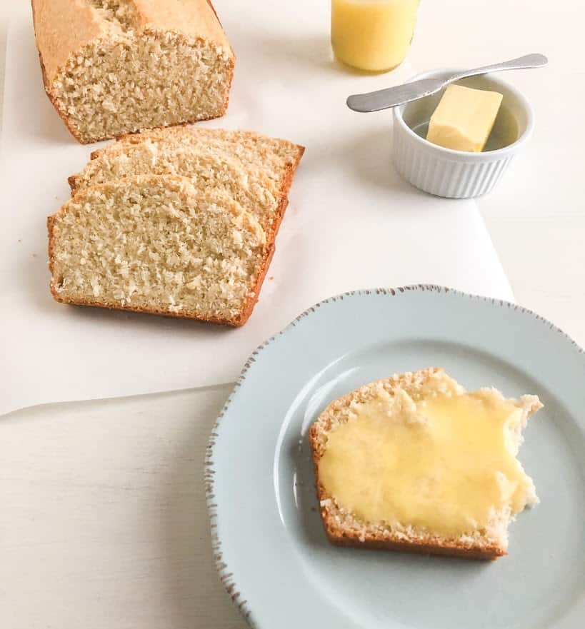 Slice of coconut bread on round light blue plate with butter and lime curd on top with bites taken out of the slice.  Sliced bread and the remaining loaf on parchment paper next to a small white round ramekin with butter and a silver butter knife. Glass jar with lime curd in the background.