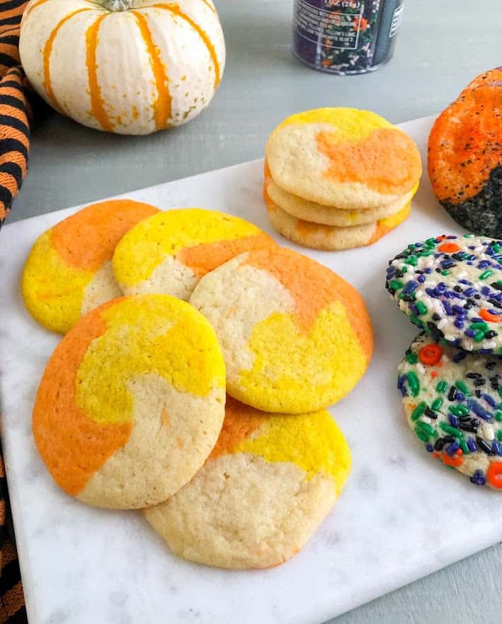 Baked sugar cookies on marble cutting board, candy corn colored sugar cookies, Halloween sprinkle sugar cookies, and sanding sugar coated sugar cookies. Black and orange striped kitchen towel and white pumpkin with orange stripes in the back ground. Jar of Halloween sprinkles also in the back ground.