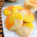 Candy corn colored sugar cookies on marble slab next to orange and black striped kitchen towel. White pumpkin with orange stripes and container of Halloween sprinkles in the back ground.
