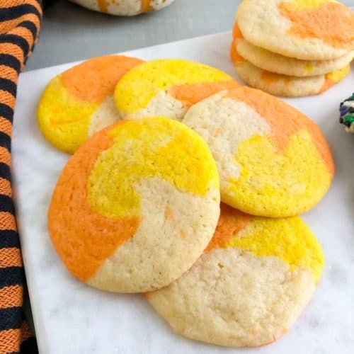 Candy corn colored sugar cookies on marble slab next to orange and black striped kitchen towel. White pumpkin with orange stripes and container of Halloween sprinkles in the back ground.