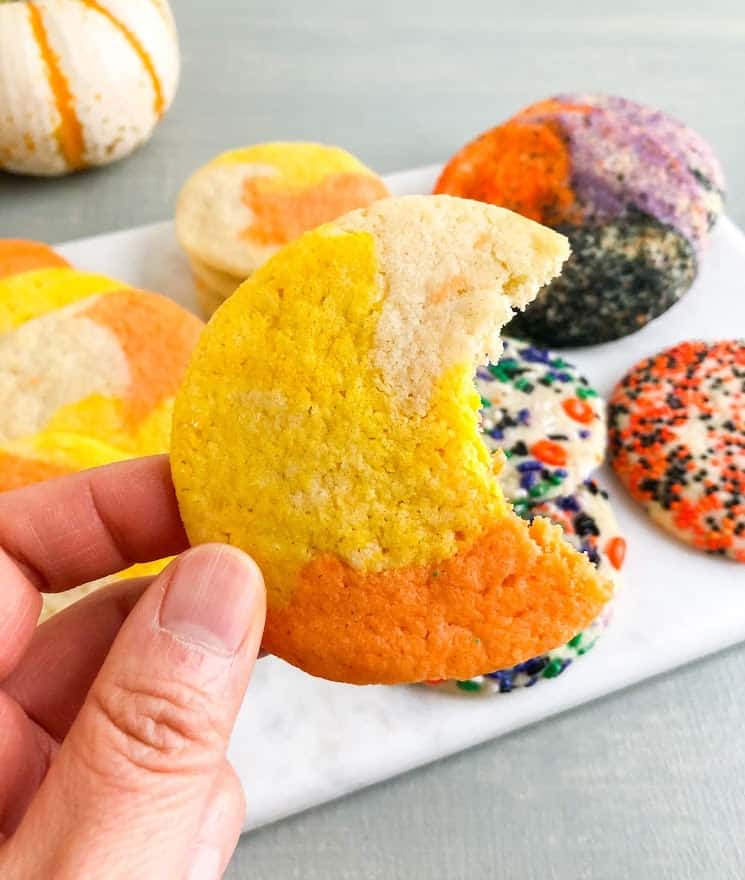 Hand holding "candy corn" colored sugar cookie with bite taken out.  Additional cookies in the back ground on marble board, some colored and other rolled in Halloween sprinkles or purple, orange, and black sanding sugar.  White pumpkin with orange stipes in the back ground.