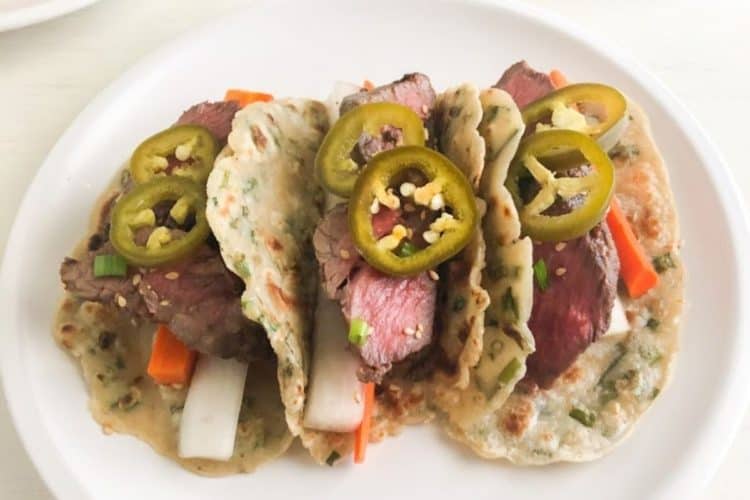 3 scallion pancake tacos on round white plate. Tacos filled with sliced short ribs, pickled daikon, carrot, and jalapenos. Small round white plates in the back ground, one with stack of scallion pancakes and the other with Korean short ribs topped with green onions.