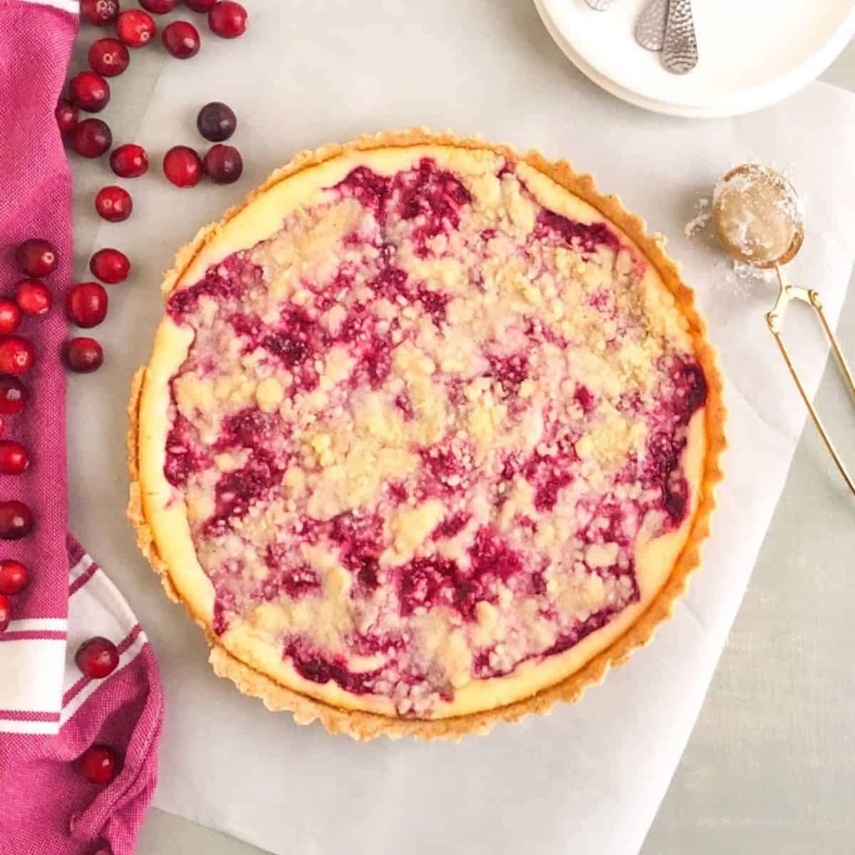 Whole baked cranberry pie on parchment paper next to pink kitchen towel with white accents and whole fresh cranberries. Small round white plates with silver cocktail forks and gold tea strainer filled with powdered sugar on the other side of the tart.