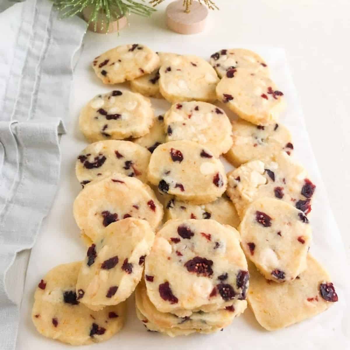 Baked cranberry shortbread cookies on parchment paper stacked and laid on top of each other next to light gray cloth napkin with green and gold glittery and wiry trees in the background.