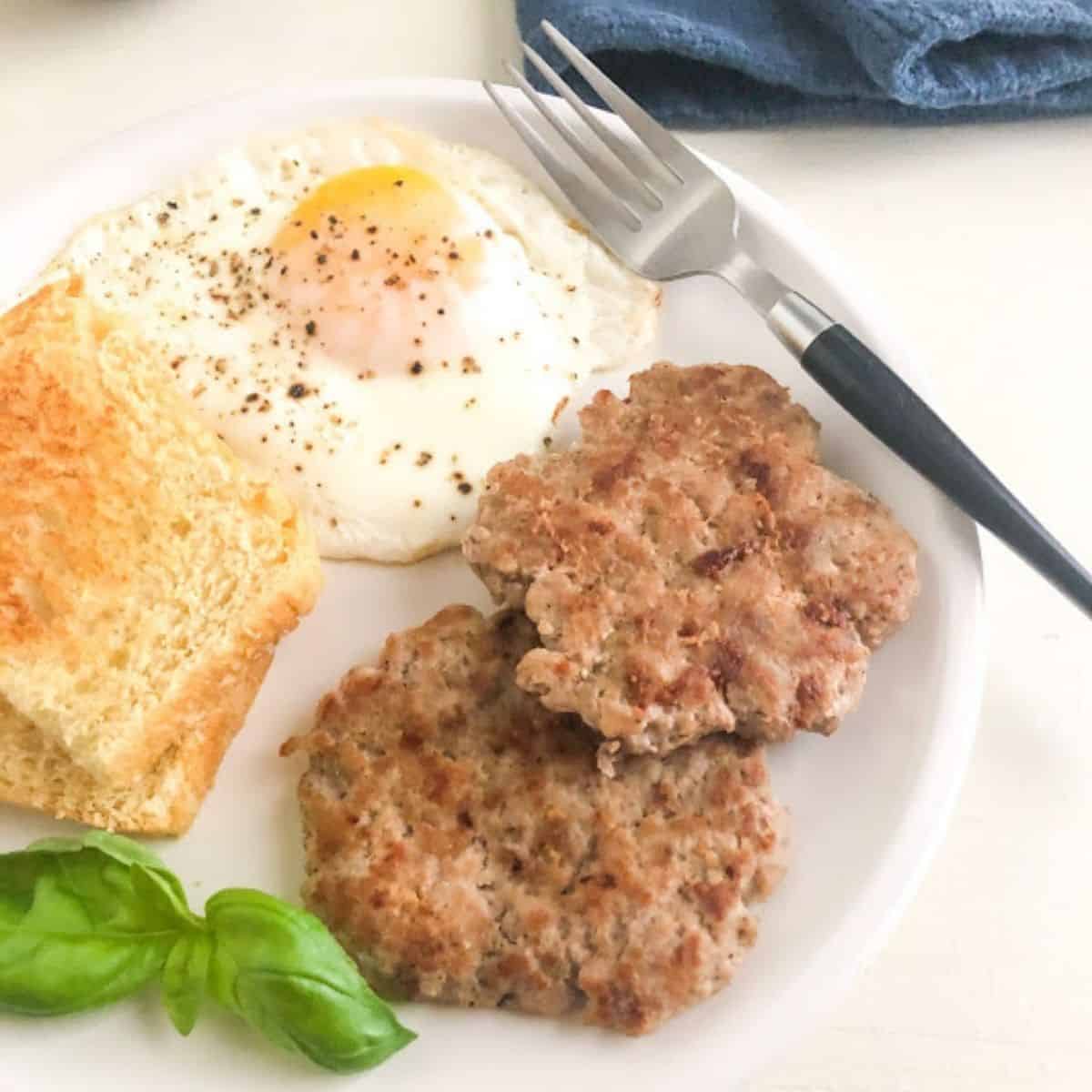 Medium round white plate with two sausage patties, over easy egg, two pieces of toast, and fresh basil as garnish and silver fork with black handle on the plate. Purple coffee cup and dark blue kitchen towel in the back ground.