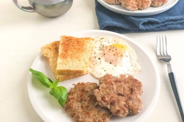 Medium round white plate with two turkey sausage patties, toast, over easy egg, and fresh basil garnish next to silver fork with black handle. Purple coffee cup net to small round white plate with additional cooked sausage on dark blue kitchen towel in the back ground.