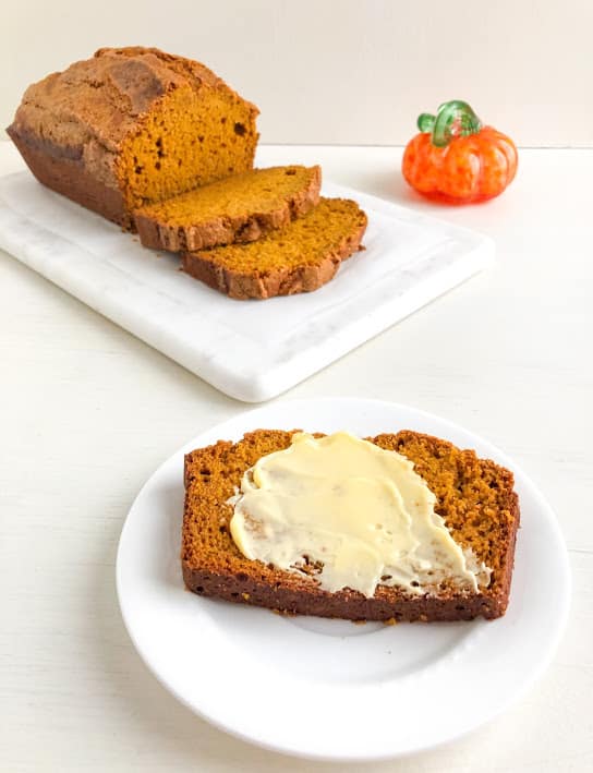 Slice of pumpkin bread with butter on small white round plate, remaining loaf and slices on white marble cutting board plus orange glass pumpkin with green stem in the background.