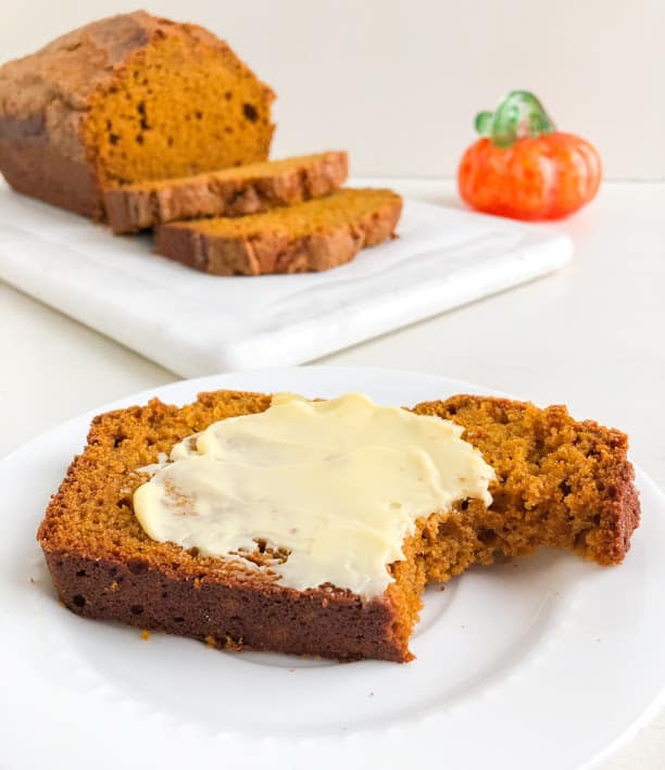 Slice of pumpkin bread with butter and bite taken out on small white round plate, remaining loaf and slices on white marble cutting board plus orange glass pumpkin with green stem in the background.