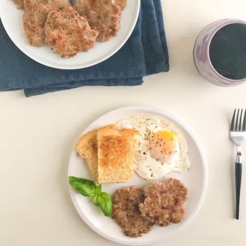 Medium round white plate with two turkey sausage patties, toast, over easy egg, and fresh basil garnish next to silver fork with black handle. Purple coffee cup net to small round white plate with additional cooked sausage on dark blue kitchen towel in the back ground.