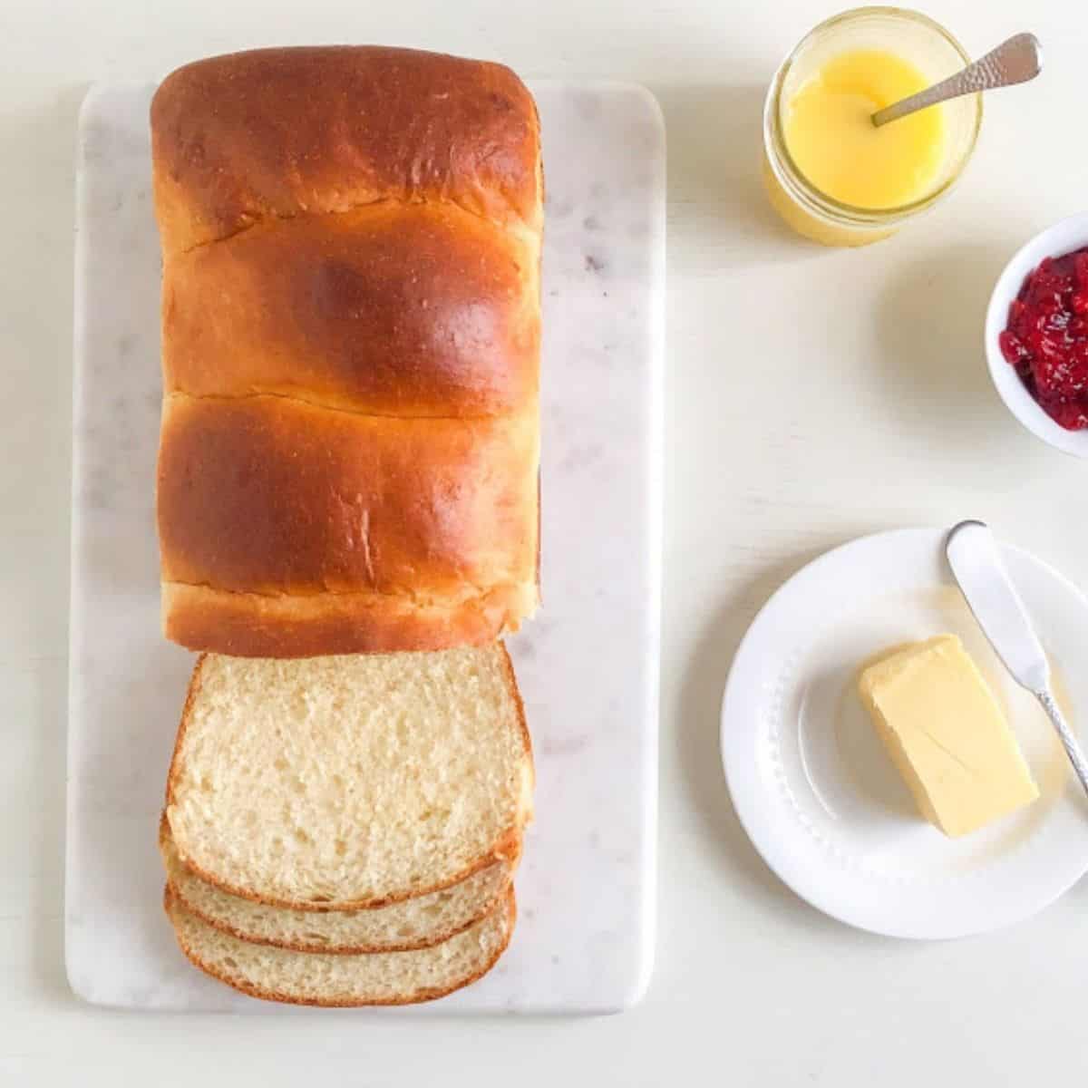 Baked bread partially sliced on marble cutting board, next to small round white plate with butter and silver butter knife. small round bowl with strawberry freezer jam, and mason jar with lemon curd and small silver spoon.