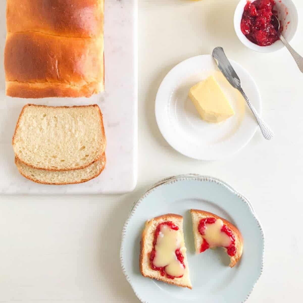 Loaf of bread partially sliced on marble cutting board, next to medium round light blue plates with a slice of bread cut in half with one bite missing and topped with jam and lemon curd. Small round while plate with butter and sliver butter knife and small round white bowl with strawberry freezer jam with small silver teaspoon in the back ground.