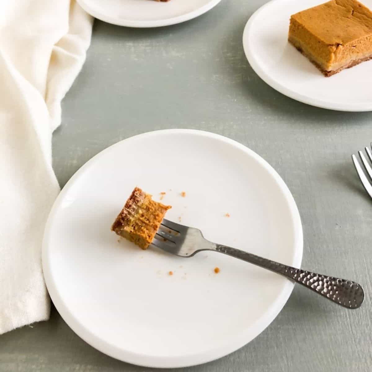 Small round white plate with silver cocktail fork with one bite of pie bar on the fork left. Additional silver cocktail forks and small round white plates with pieces of pie bars in the background. Cream cloth napkin next to plates.