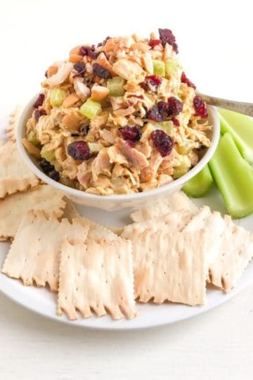 Medium white round plate with plain crackers and celery sticks with small bowl of chicken salad in the middle. Extra cranberries and cashews on top of the chicken salad with a small silver cocktail spoon.