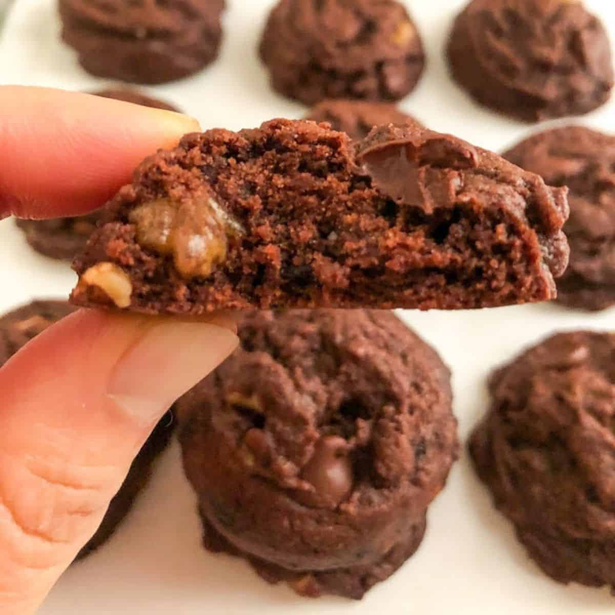 Hand holding brownie cookie with bite taken out of it.  Additional cookies in the background on parchment paper.