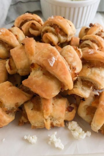 Rugelach arranged on top of each other on parchment paper with crumbles of feta. Light gray linen napkin and small round white ramekin in the background.