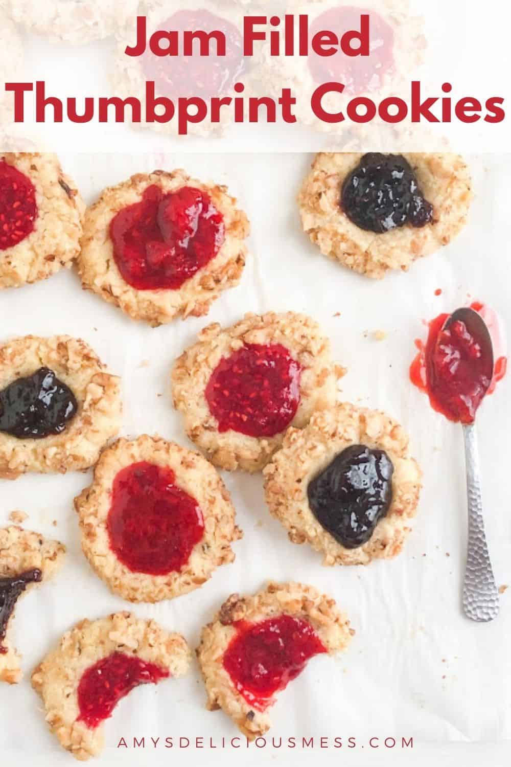 Thumbprint cookies baked and filled with strawberry, raspberry, or black forest cherry jam. Arranged on parchment with bites taken out of 3 coolies with silver cocktail spoon with additional jam next to the cookies.