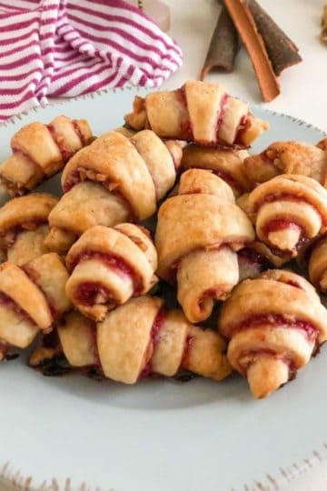 Small round light blue plate with stack of baked cookies arranged on each other. Pink and white stripped kitchen towel, cinnamon sticks, and walnuts in the background for decoration.